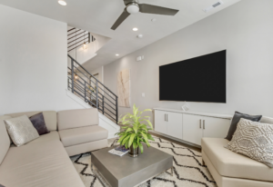 Modern living room with beige sectional sofa, large TV, plant on a gray table, and a staircase with a metal railing.